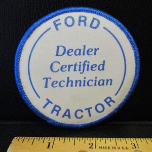 Op de afbeelding: Een ronde blauwe en witte patch met de tekst "FORD TRACTOR" in een blauwe cirkel. De tekst "Dealer Certified Technician" is in zwart binnen de cirkel gedrukt.