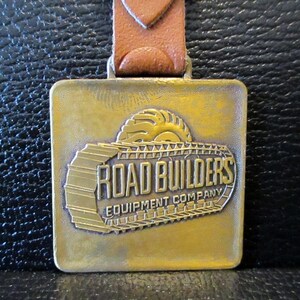 May include: A gold-colored metal keychain with a brown leather strap. The keychain features a raised image of a bulldozer with the text "Road Builders Equipment Company".