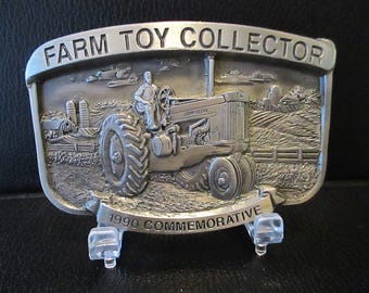 May include: A silver-toned metal belt buckle with the words "FARM TOY COLLECTOR" across the top. The buckle features a tractor with a driver, a farm scene, and the text "1990 COMMEMORATIVE".