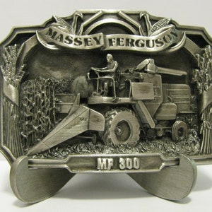 May include: A silver-toned belt buckle featuring a detailed 3D image of a Massey Ferguson MF 300 combine harvester in a field of wheat. The buckle has a banner with the words "Massey Ferguson" and a raised design of wheat stalks.