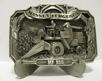 May include: A silver-toned belt buckle featuring a detailed 3D image of a Massey Ferguson MF 300 combine harvester in a field of wheat. The buckle has a banner with the words "Massey Ferguson" and a raised design of wheat stalks.
