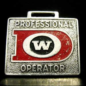Op de afbeelding: Een zilveren metalen sleutelhanger met een rode en witte cirkel met de letter "W" in zwart. De sleutelhanger heeft de tekst "Professional Operator" boven en onder de cirkel.