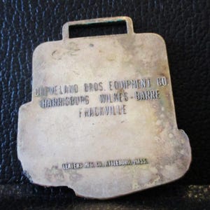 May include: A silver-coloured metal tag in the shape of a briefcase, with a rectangular hole at the top. The tag is engraved with the text "CLEVELAND BROS. EQUIPMENT CO HARRISBURG WILKES-BARRE FRACKVILLE" and "LEAVENS MFG. CO. ATTLEBORO, MASS".