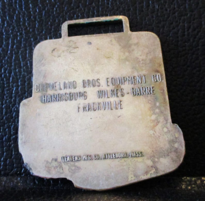May include: A silver-coloured metal tag in the shape of a briefcase, with a rectangular hole at the top. The tag is engraved with the text "CLEVELAND BROS. EQUIPMENT CO HARRISBURG WILKES-BARRE FRACKVILLE" and "LEAVENS MFG. CO. ATTLEBORO, MASS".