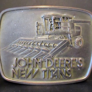 May include: A rectangular metal belt buckle with a raised relief of a John Deere combine harvester. The buckle has the words "JOHN DEERE'S NEW TITANS" embossed on the front. The buckle has a silver finish.