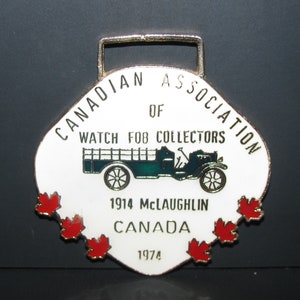 Puede incluir: Un llavero de reloj de esmalte blanco con un borde dorado y un diseño de bandera canadiense rojo, blanco y azul. El llavero presenta un automóvil McLaughlin verde de 1914 y el texto "Canadian Association of Watch Fob Collectors 1914 McLaughlin Canada 1974".
