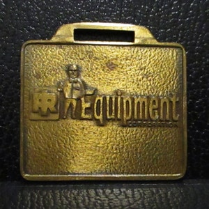 Puede incluir: Un llavero de metal dorado con un logotipo de RR Equipment Corporation. El logotipo presenta a un hombre de dibujos animados sosteniendo una llave inglesa y el texto "RR Equipment Corporation".