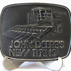 May include: A silver metal belt buckle featuring a John Deere combine harvester with the text "JOHN DEERE'S NEW TITANS".