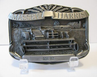 May include: A silver-toned belt buckle featuring a detailed depiction of a Massey Harris combine harvester in a field setting. The buckle also includes the text "MASSEY HARRIS" and "MH 92".