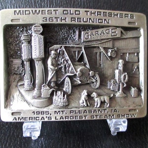 Può includere: Una fibbia per cintura in argento con un'immagine 3D di una scena di stazione di servizio vintage. La fibbia raffigura un'auto, pompe di benzina, un garage e due cani. Il testo "MIDWEST OLD THRESHERS 36TH REUNION GARAGE AUTO SERVICE 1985, MT. PLEASANT, IA. AMERICA'S LARGEST STEAM SHOW" è inciso sulla fibbia.