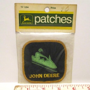 May include: A John Deere patch featuring a green snowmobile design on a black background, with gold trim. The patch is in a clear plastic package with the word "patches" and the John Deere logo.
