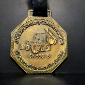 Puede incluir: Un medallón conmemorativo de metal dorado con una correa de cuero negro. El medallón presenta una retroexcavadora con el texto "J.I. Case Company Commemorative 15 Years of Backhoe Production CK 580 B"