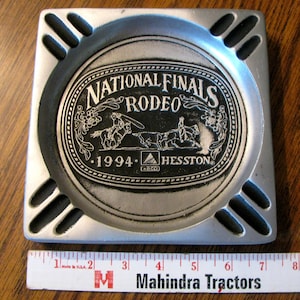 May include: A square, silver ashtray with a black interior and four cigarette rests. The centre features the text "NATIONAL FINALS RODEO 1994 HESSTON" with rodeo illustrations. The ashtray is approximately 13 cm square.
