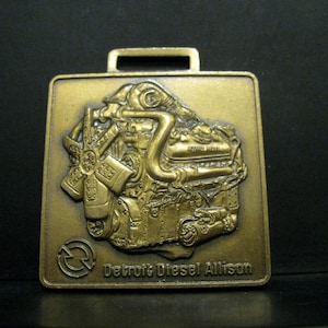 May include: A gold-colored metal keychain featuring a detailed relief of a Detroit Diesel Allison engine. The keychain has a circular arrow symbol and the text "Detroit Diesel Allison".