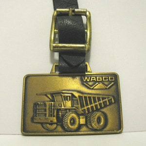May include: A gold-colored metal watch fob with a black leather strap and a buckle. The fob features a raised image of a dump truck with the text "WABCO" above it.