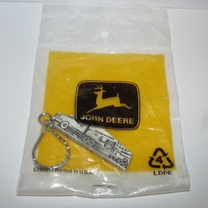 May include: A silver John Deere keychain featuring a detailed tractor design. The keychain is packaged in a clear plastic bag with a yellow background and the John Deere logo. The bag also includes the text "E388617 Printed in U.S.A." and a recycling symbol.