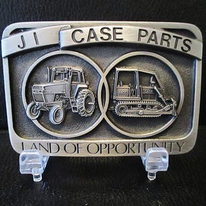 Può includere: Fibbia per cintura in argento con un trattore e un bulldozer in rilievo, circondati da un cerchio con il testo "JI CASE PARTS LAND OF OPPORTUNITY".
