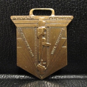 May include: A gold-coloured metal pocket watch fob with the text "Shull Perforator Knife Perforator Side View of Tong" engraved on it.