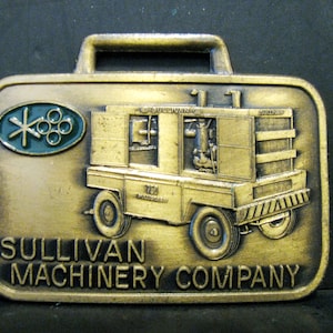Puede incluir: Una hebilla de cinturón de metal de color dorado con una imagen detallada de un pequeño vehículo de construcción amarillo con el texto "Sullivan Machinery Company" y un logotipo verde con una estrella y círculos.