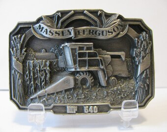 May include: A silver-toned metal belt buckle featuring a detailed 3D image of a Massey Ferguson MF 540 combine harvester in a field of wheat. The buckle has a banner with the words "Massey Ferguson" and the words "MF 540" are engraved on the bottom.