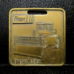 Op de afbeelding: Een gouden metalen sleutelhanger met een Rex Trashmaster bulldozer. De sleutelhanger heeft een gat om aan een sleutelring te bevestigen. De bulldozer is in zijaanzicht afgebeeld met de tekst "Rex" en "Trashmaster" op de sleutelhanger.