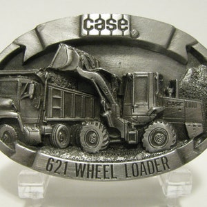 Könnte beinhalten: Eine silberne Gürtelschnalle mit einem 3D-Design eines Case 621 Radladers und eines Muldenkippers. Die Schnalle trägt das Case-Logo und den Text "621 Wheel Loader".
