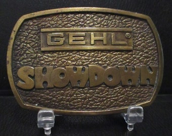 May include: A brass belt buckle with a textured surface. The buckle features the words "GEHL" and "SHOWDOWN" in raised lettering.