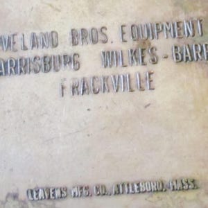 May include: Close-up of a weathered metal surface with embossed text. The text reads "CLEVELAND BROS. EQUIPMENT CO HARRISBURG WILKES-BARRE FRACKVILLE" and "LEAVENS MFG. CO., ATTLEBORO, MASS."