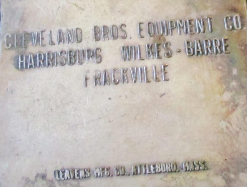 May include: Close-up of a weathered metal surface with embossed text. The text reads "CLEVELAND BROS. EQUIPMENT CO HARRISBURG WILKES-BARRE FRACKVILLE" and "LEAVENS MFG. CO., ATTLEBORO, MASS."
