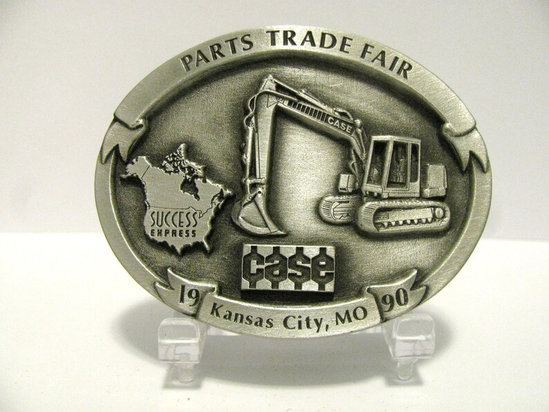 Puede incluir: Una hebilla de cintur&oacute;n plateada con una excavadora Case, el logotipo de Case y el texto "Parts Trade Fair 19 Kansas City, MO 90". La hebilla tambi&eacute;n incluye un mapa de Am&eacute;rica del Norte con el texto "Success Express".