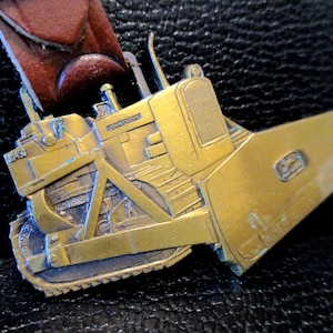 Op de afbeelding: Een gouden metalen gesp met een gedetailleerd model van een John Deere D450 bulldozer. De bulldozer heeft een opgeheven blad en een getextureerd rooster aan de voorkant.