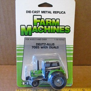 May include: A green and black Deutz-Allis 7085 tractor with dual wheels, a die-cast metal replica, in a plastic package. The package says "Farm Machines" and "1/64 Scale".