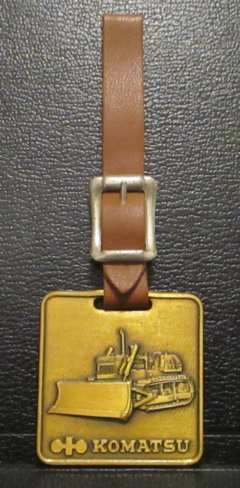 May include: A gold-coloured metal keychain with a brown leather strap and a silver buckle. The keychain features a detailed image of a Komatsu bulldozer with the text "Komatsu" below it.
