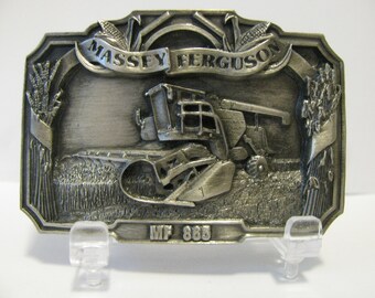 May include: A silver-toned metal belt buckle featuring a detailed image of a Massey Ferguson MF 885 combine harvester in a field of wheat. The buckle has a banner with the words "Massey Ferguson" and the words "MF 885" are engraved below the image.