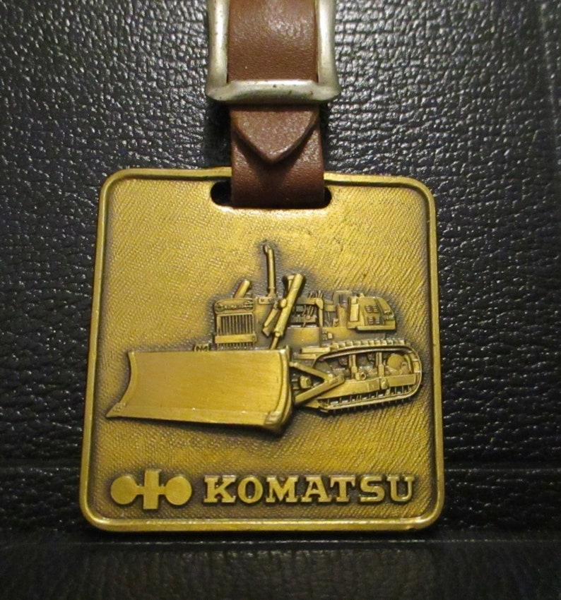 May include: A gold-coloured metal keychain featuring a Komatsu bulldozer with a brown leather strap. The keychain has the Komatsu logo and the words "KOMATSU" on it.