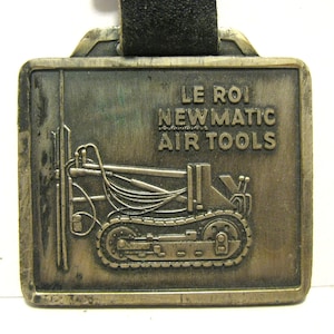 May include: A gold-colored metal keychain with a black leather strap. The keychain features a small, detailed image of a construction vehicle with the text "LE ROI NEWMATIC AIR TOOLS" above it.