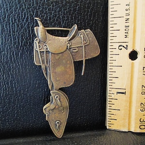 May include: A bronze-colored metal saddle pin brooch. The saddle is detailed with stitching and a stirrup. The pin is approximately 2 inches in length.