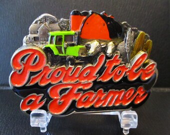 May include: A silver belt buckle with a black background and red lettering that says "Proud to be a Farmer". The buckle features a green tractor, a red barn, and cows.