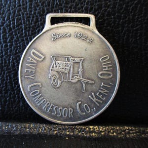 Puede incluir: Un llavero de reloj de bolsillo de metal plateado con un diseño circular. El llavero presenta una pequeña imagen de un camión con el texto "Davey Compressor Co., Kent, Ohio" y "Since 1922".
