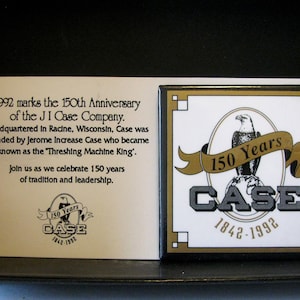 Puede incluir: Una placa blanca con bordes dorados que conmemora el 150 aniversario de la J.I. Case Company. La placa presenta el texto "1992 marks the 150th Anniversary of the J.I. Case Company. Headquartered in Racine, Wisconsin, Case was founded by Jerome Increase Case who became known as the 'Threshing Machine King'. Join us as we celebrate 150 years of tradition and leadership." Una placa más pequeña con bordes dorados presenta un águila calva, una pancarta con el texto "150 Years", y el texto "CASE 1842-1992".