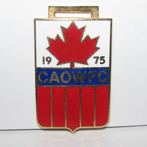 Op de afbeelding: Gouden metalen pin met een rood esdoornblad en blauwe en witte strepen. De pin heeft de tekst "CAOWFC" en "1975".