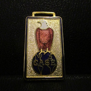 Puede incluir: Fob de reloj de metal dorado con un águila de esmalte rojo, blanco y azul de pie sobre un globo azul con la palabra "CASE" en letras doradas.