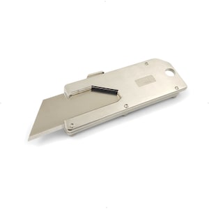 May include: A silver utility knife with a retractable blade. The knife has a rectangular body with rounded edges and a small hole for a lanyard. The blade is extended and ready for use. The text "CATO" is visible on the body of the knife.