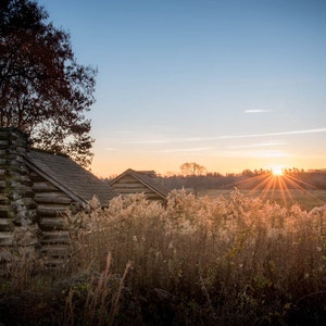 May include: A rustic wooden cabin with a stone chimney stands in a field of tall grass, bathed in the golden light of a setting sun. The sun's rays create a starburst effect in the sky.