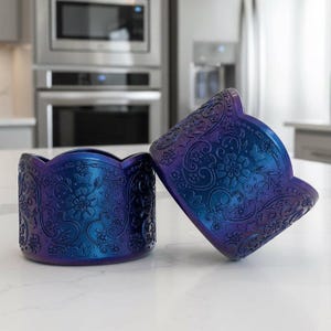 May include: Two iridescent blue and purple decorative containers with a floral and scrollwork design. The containers have a scalloped edge and are made of a shiny material. One container is upright, and the other is tilted.