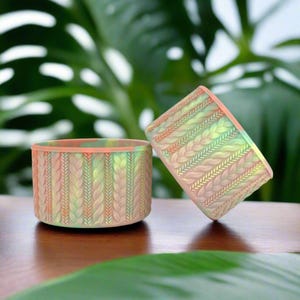May include: Two small, round, ceramic planters with a braided pattern in shades of pink, green, and yellow. The planters are on a wooden surface.
