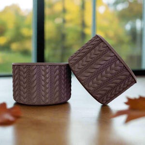 May include: Two brown, textured planters with a cable knit design. One planter is upright, while the other is tilted, showcasing the intricate pattern. The planters are made of a solid material and are set on a wooden surface, with a blurred background.