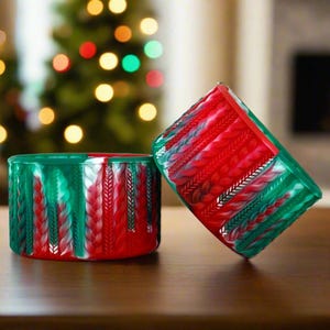 May include: Two red and green Christmas-themed candle holders with a braided pattern. The holders are made of a textured material and have a white and silver swirl design.