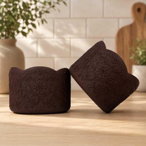 May include: Two dark brown cylindrical containers featuring a scalloped top edge and an intricate embossed floral pattern with swirling vines and small blossoms covering the entire exterior surface of each piece, displayed on a wooden surface.