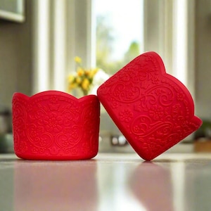 Puede incluir: Dos moldes para cupcakes de silicona roja con un patrón floral. Los moldes son de silicona flexible y tienen un borde festoneado.